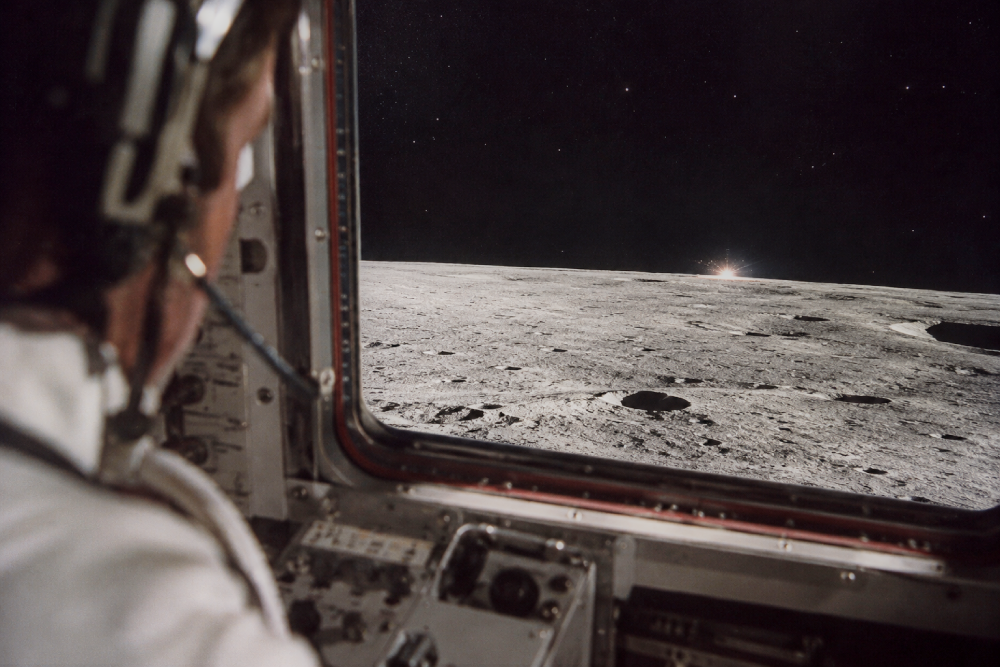 An astronaut viewed from behind looks through a spacecraft window at the cratered lunar surface.