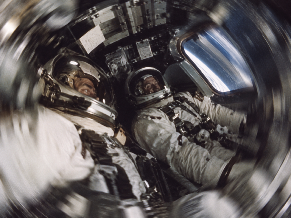 Two astronauts in pressurized spacesuits inside a spacecraft, with the Earth visible through a window and a blurred, spinning effect around the frame.