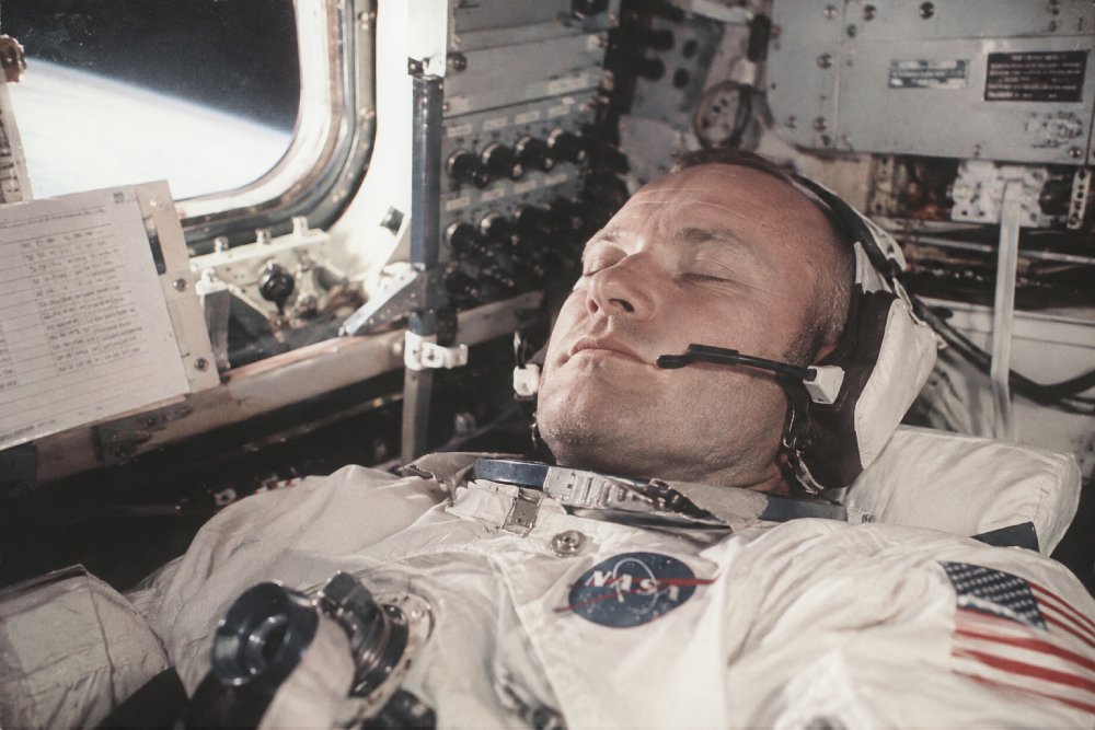 Astronaut sleeping in a spacecraft, wearing a headset and NASA flight suit, with the curve of the Earth visible through a window.