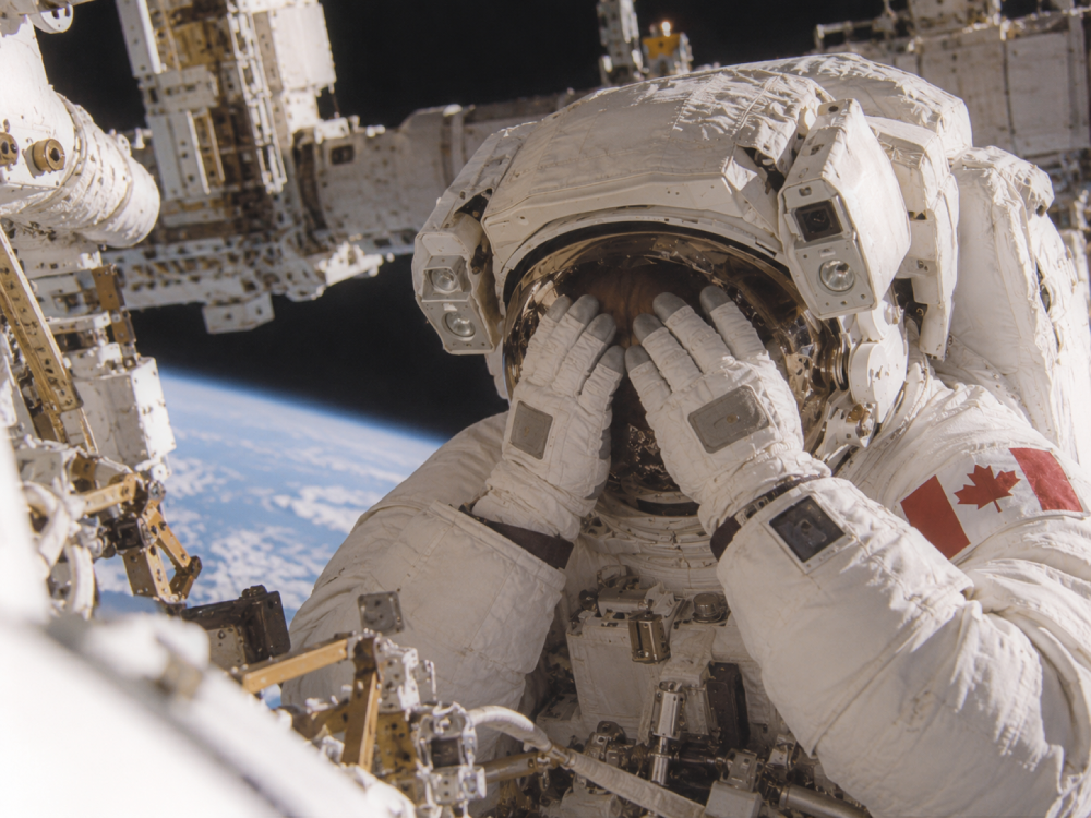 An astronaut in a spacesuit with a Canadian flag patch covers their face with gloved hands while working outside the International Space Station above Earth.
