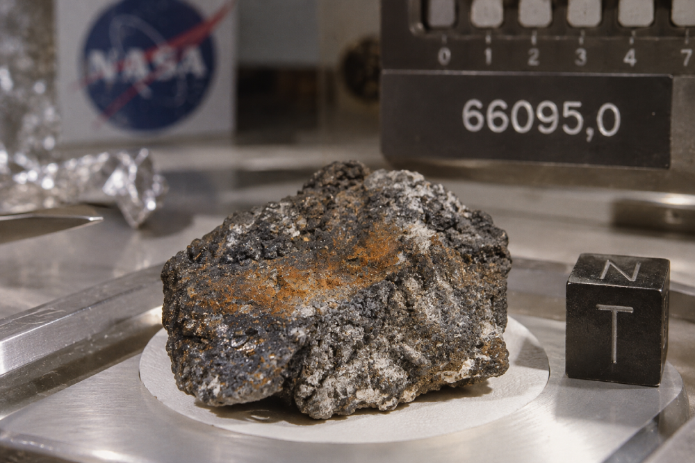 A lunar rock sample labeled “66095,0” sitting on a scale next to a small cube labeled “NT,” with a NASA logo in the background.