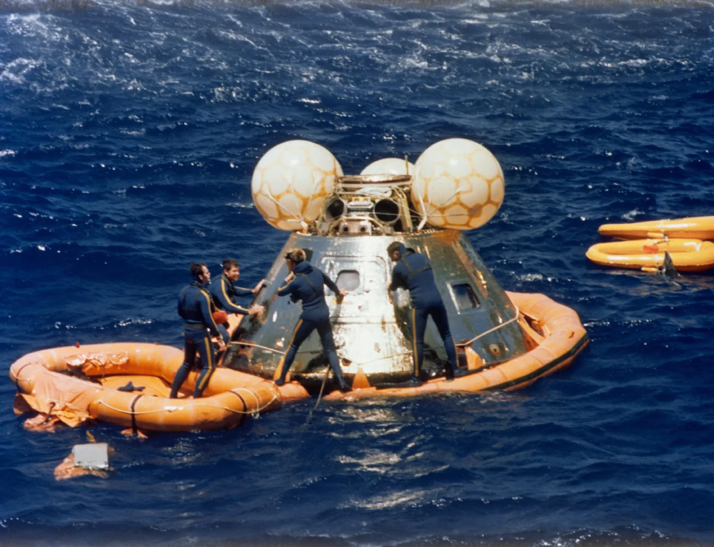 Three divers in dark wetsuits assist the Apollo command module floating in the ocean, surrounded by orange flotation collars.