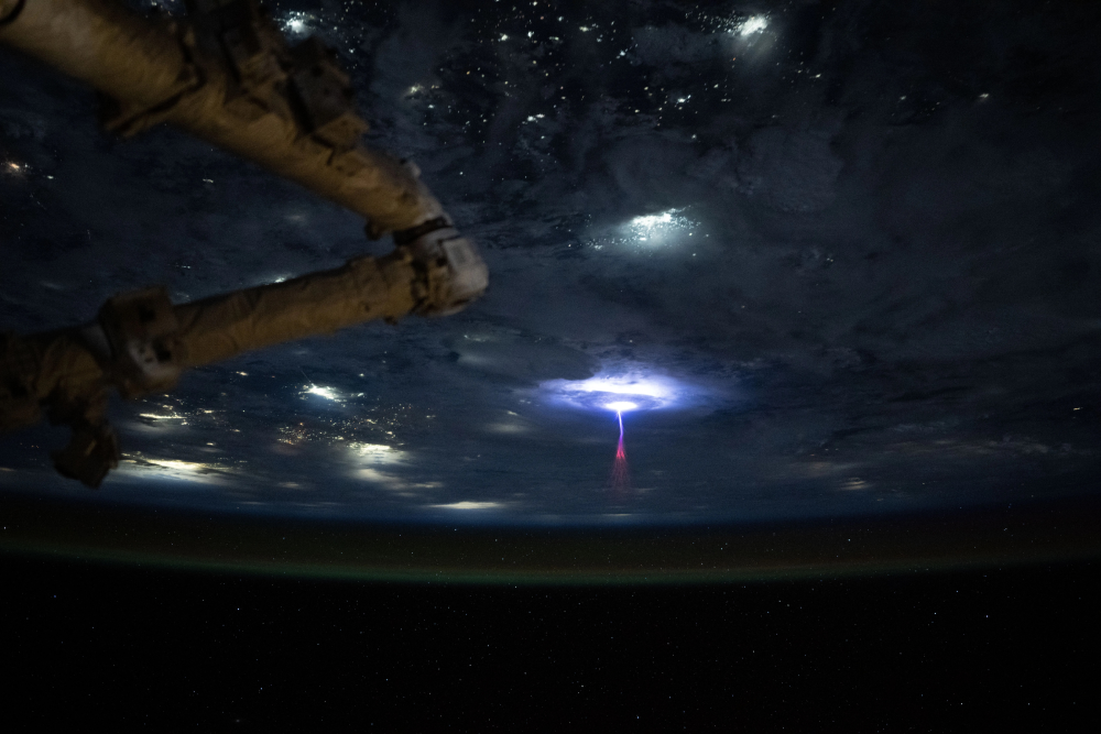 A red sprite lightning discharge illuminates clouds above Earth, viewed from the International Space Station.