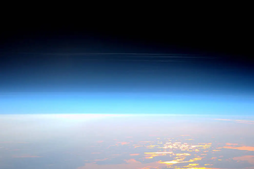 Earth’s horizon and thin blue atmosphere viewed from space, with sunlight reflecting off clouds below.