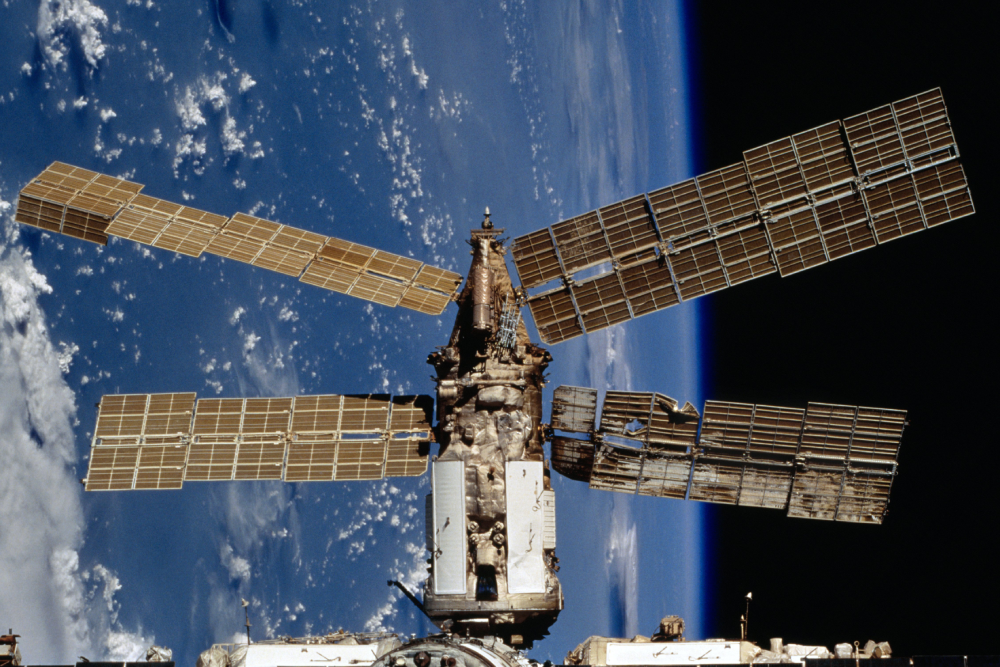 The Mir space station orbits Earth with its solar arrays extended against the backdrop of blue clouds and the dark curve of space.