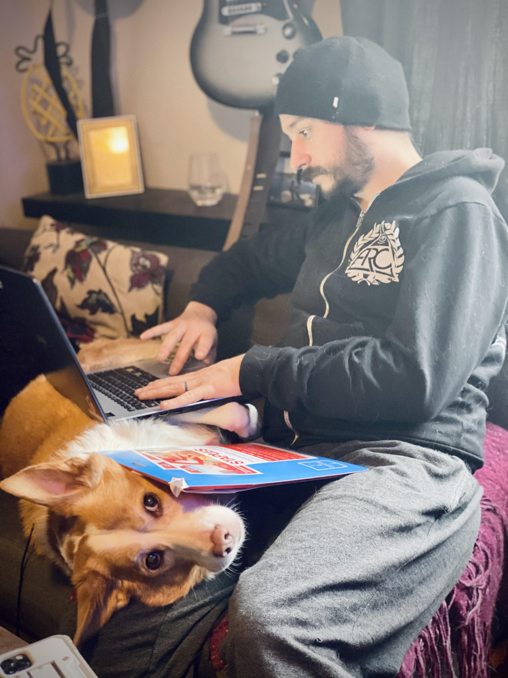 A person wearing a beanie and black hoodie sits on a couch working on a laptop, with a dog resting on their lap while balancing a notebook on its head.