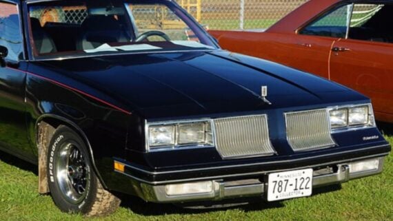 Black Oldsmobile Cutlass parked on grass with a Minnesota collector license plate reading 787-122.