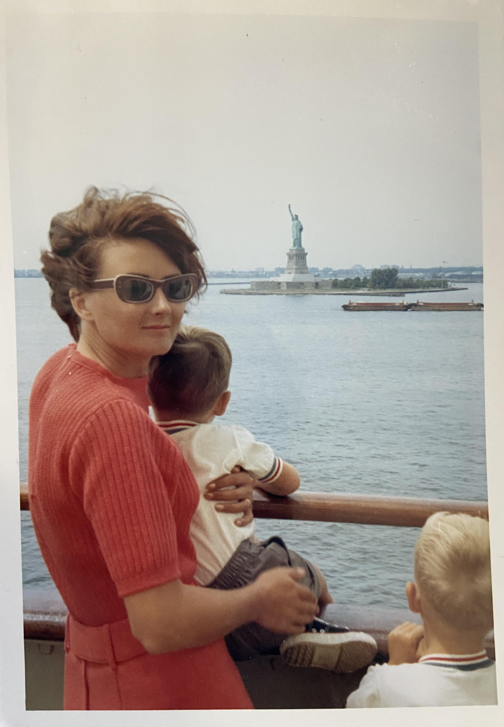 A woman in a red top and sunglasses holds a young child while standing on a boat deck with another child, looking out toward the Statue of Liberty in the distance.