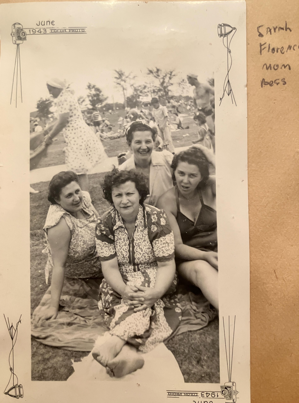 Four women sitting together on a grassy lawn with other people picnicking in the background in a 1943 photograph.