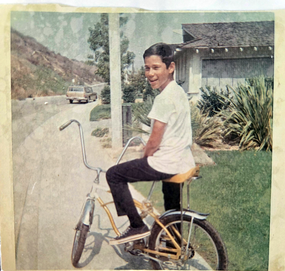 A young boy in a white t-shirt and dark pants sits on a bicycle on a residential street.