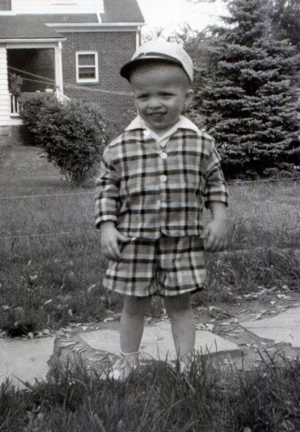 A young child wearing a plaid jacket and shorts with a cap stands in a yard in front of a house.