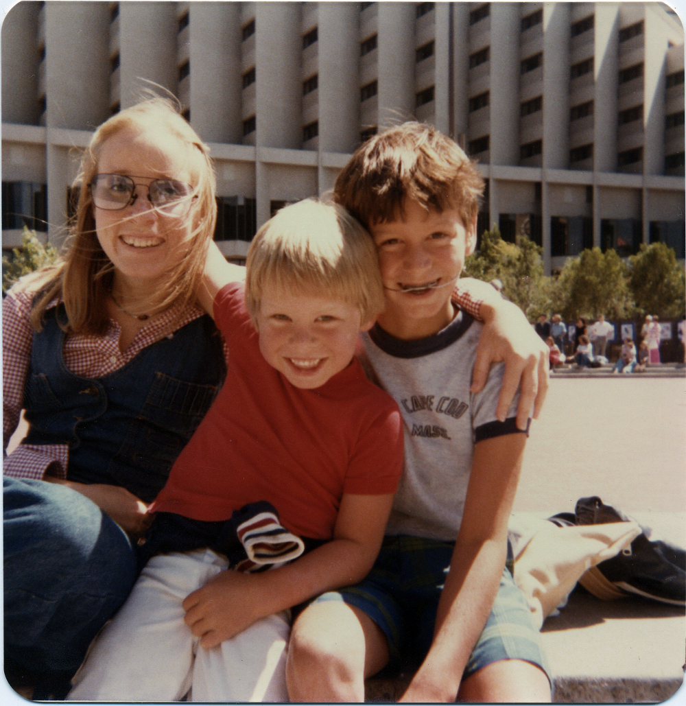 A young woman with an arm around two smiling boys, one wearing a headgear orthodontic appliance, sitting in front of a large concrete building.
