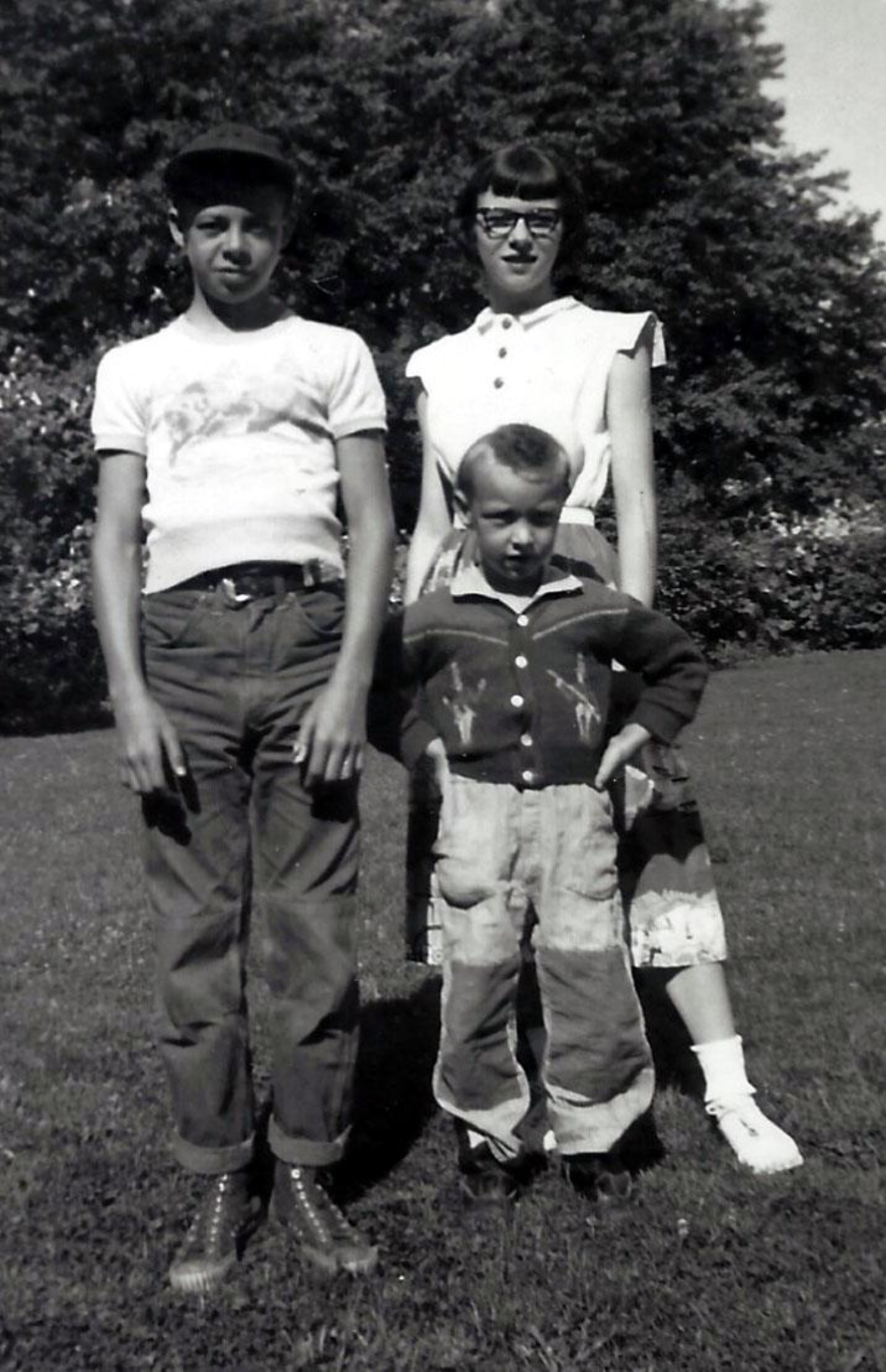 A black and white photograph of an older boy, a young woman, and a young boy standing together in a grassy yard.