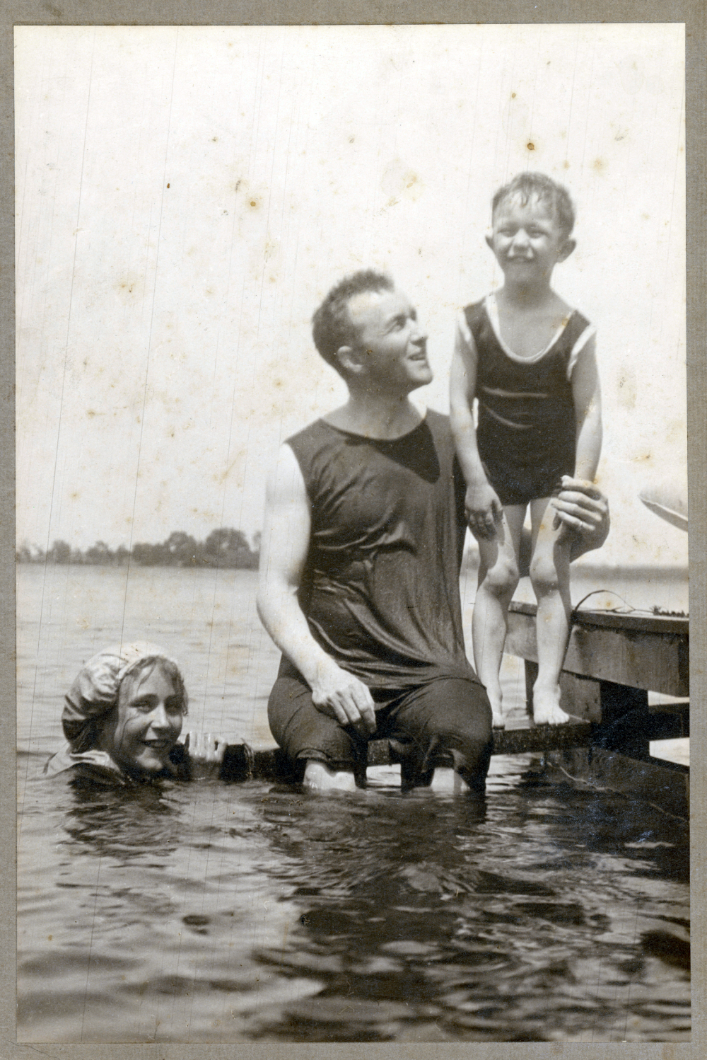A woman in a swim cap is in the water, while a man sits on a dock and a smiling child stands beside him in a swimsuit. It appears to be a vintage lakeside scene.