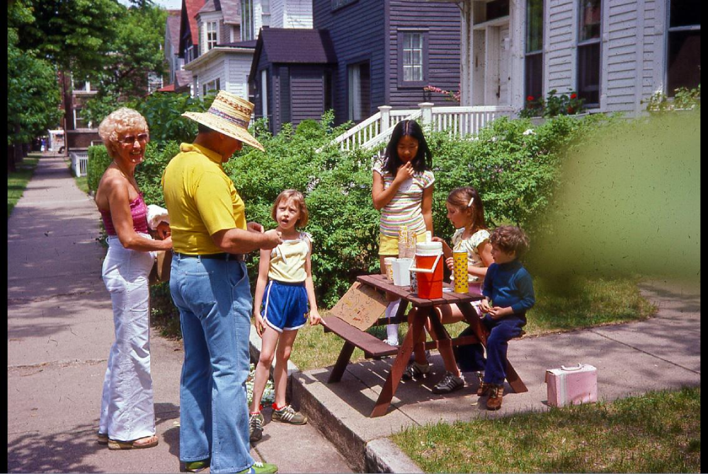 A man in a straw hat and three children gather at a small wooden picnic table on a sidewalk, with a woman standing nearby.
