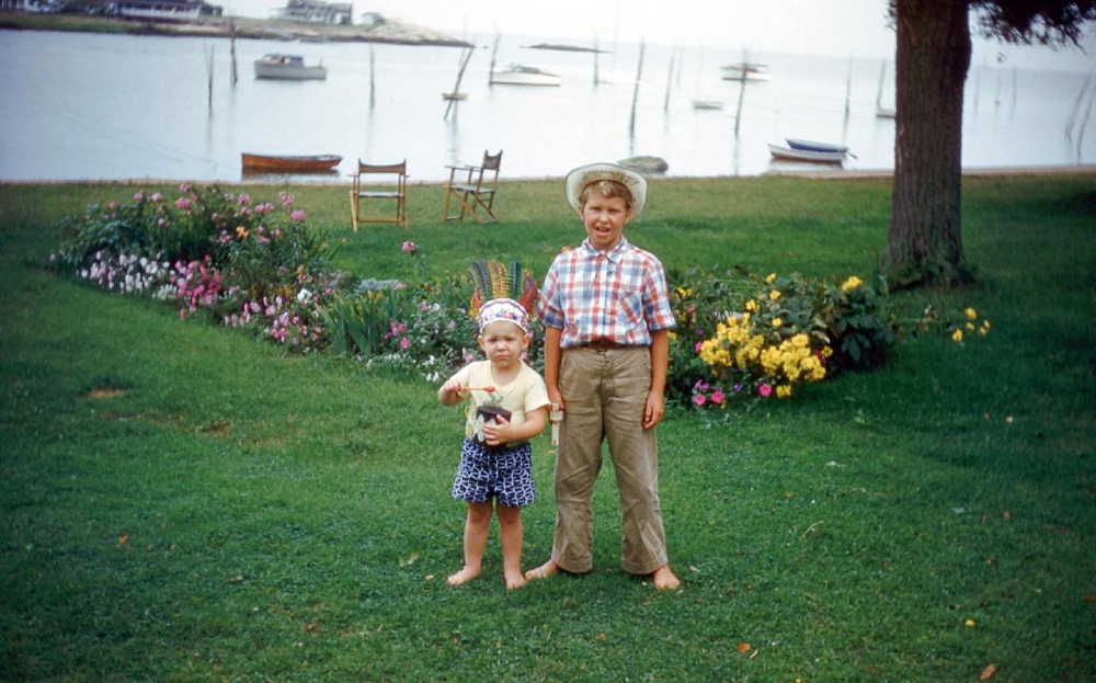 Two boys, one wearing a feathered headband, standing barefoot on a lawn in front of a bay with small boats.