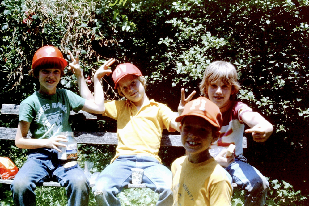 Four children wearing baseball caps sit together on a wooden bench outdoors, making peace signs and smiling at the camera.