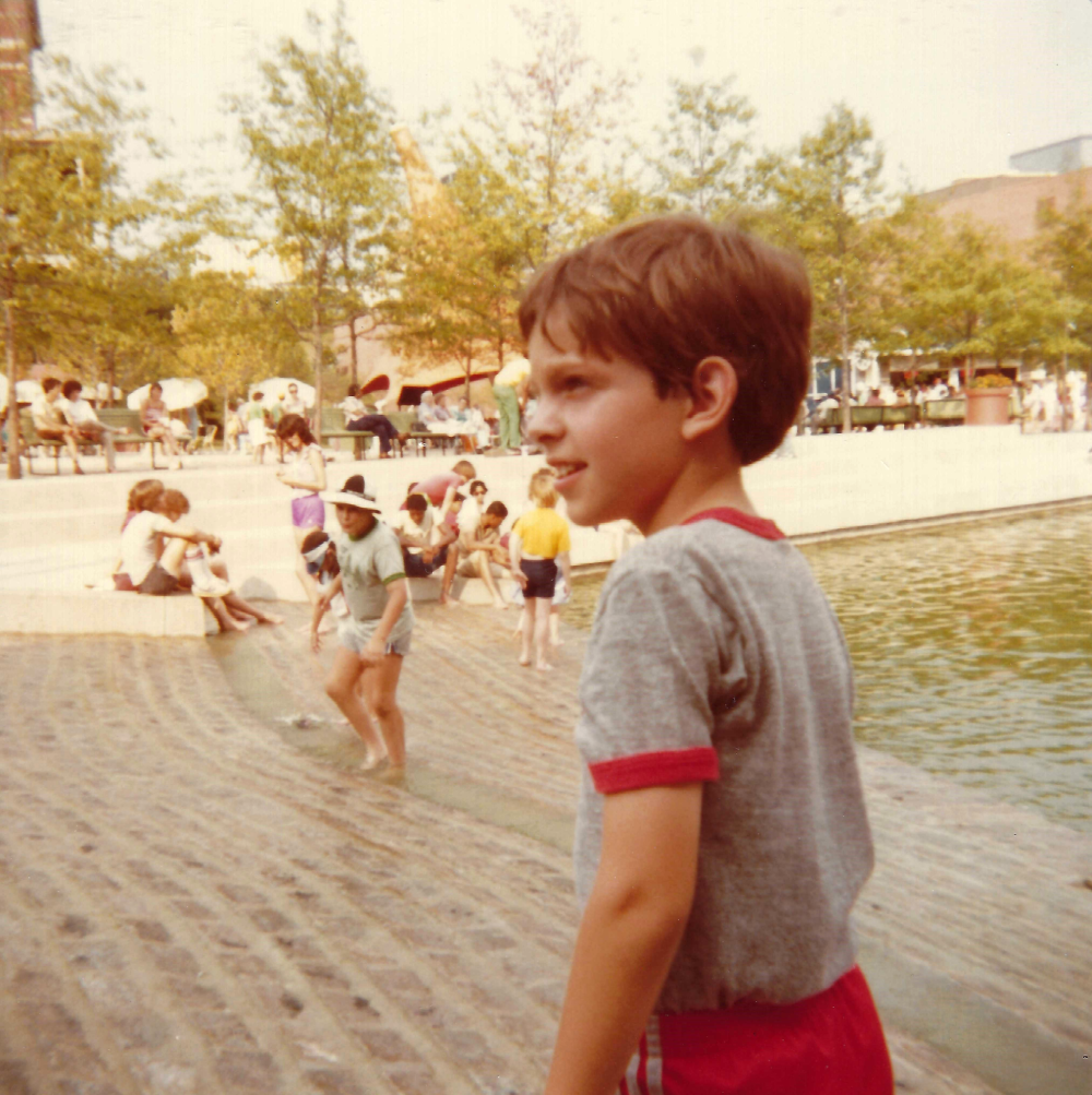 A boy in a gray shirt and red shorts stands in the foreground at a park with a wading pool where other children are playing.