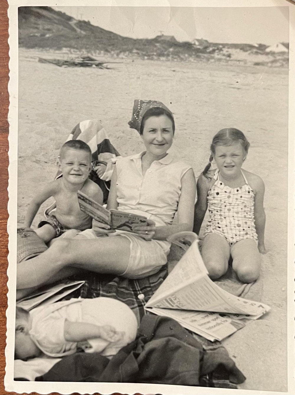 A woman sits on a sandy beach reading a book to a young boy and girl, with a baby lying on a blanket in the foreground.