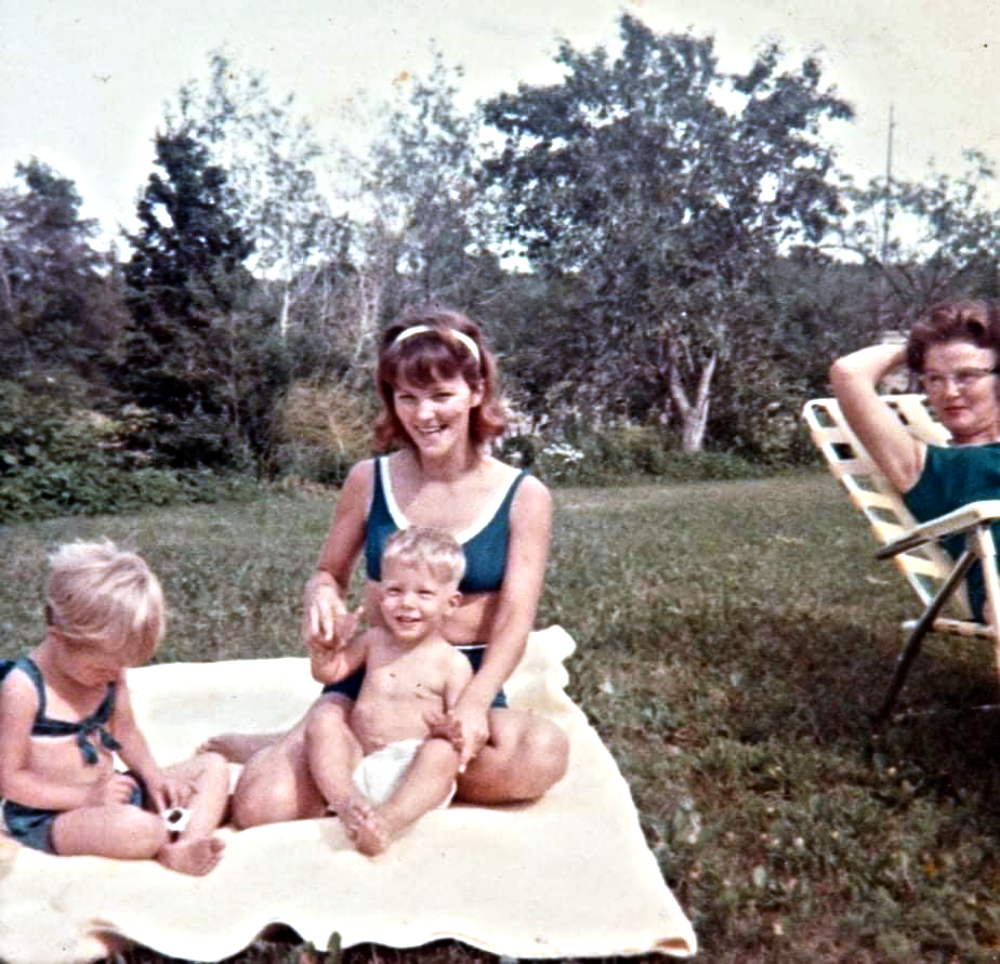 A woman in a swimsuit sits on a blanket with two young children, while another woman sits in a lawn chair nearby outdoors.