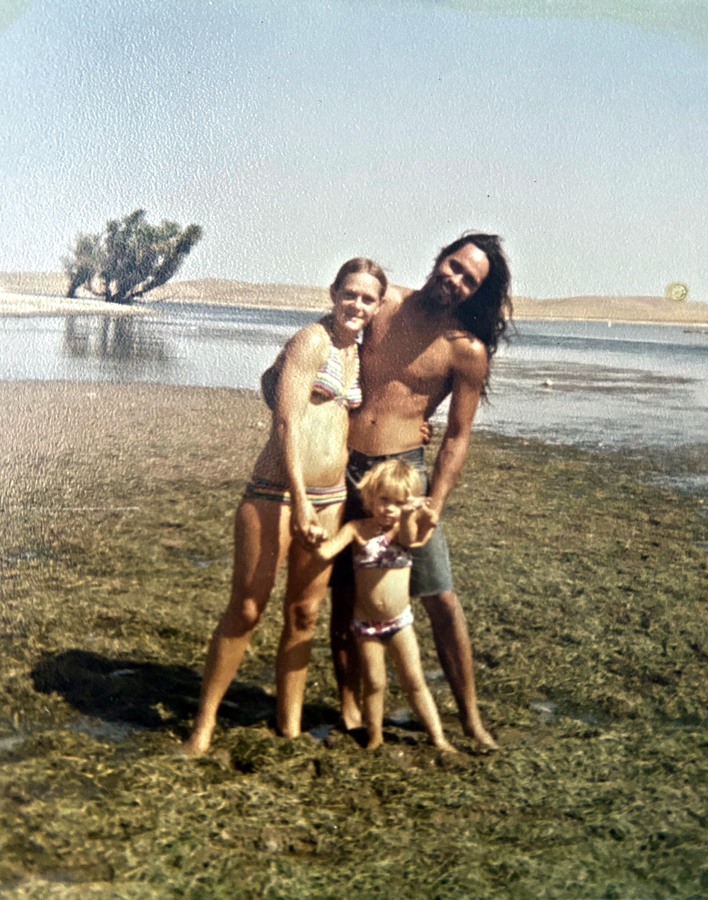 A man, woman, and young child stand together in the shallow water at a beach.