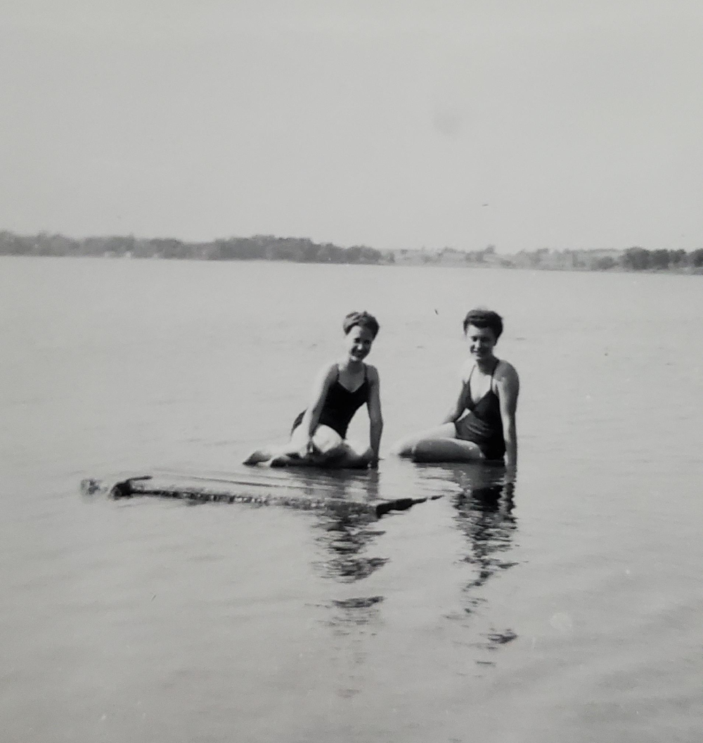 Two women in vintage swimsuits sitting on a floating wooden platform in a lake.