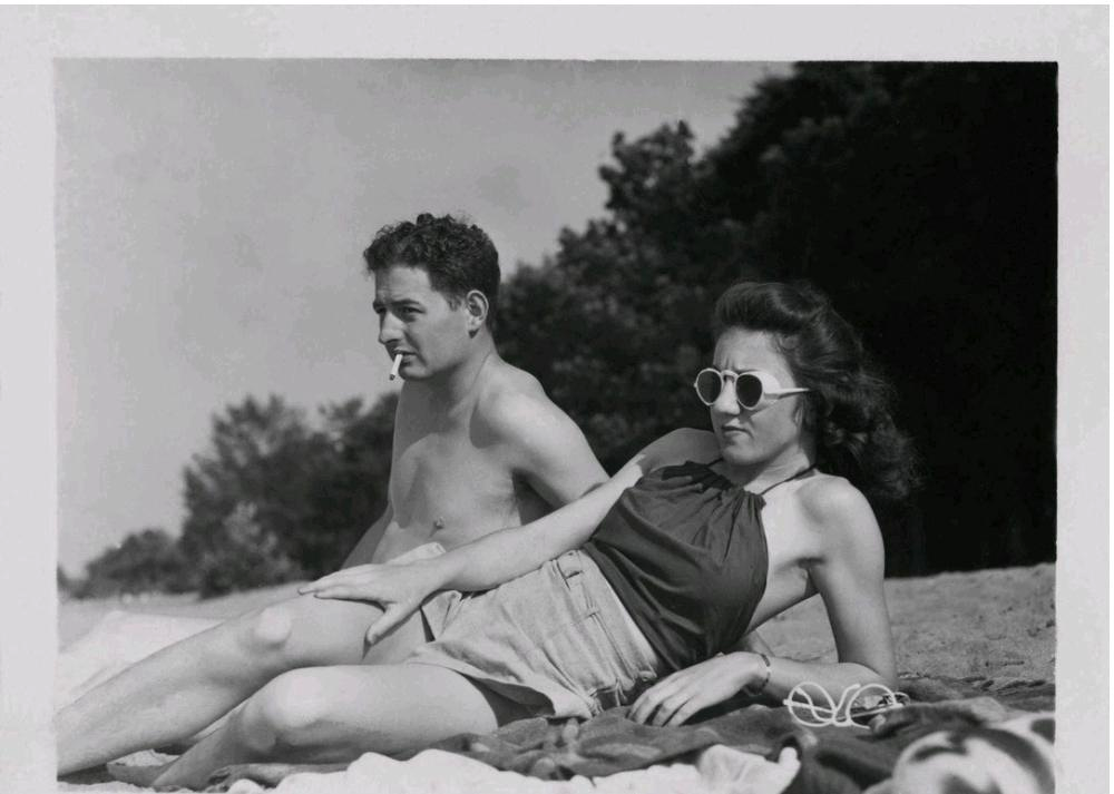 A man with a cigarette in his mouth and a woman wearing round sunglasses lying on a beach towel.