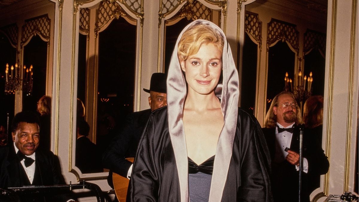 A woman in a black dress with a satin hood stands smiling at a formal event. People in tuxedos, elegant mirrors, and chandeliers are visible in the background.