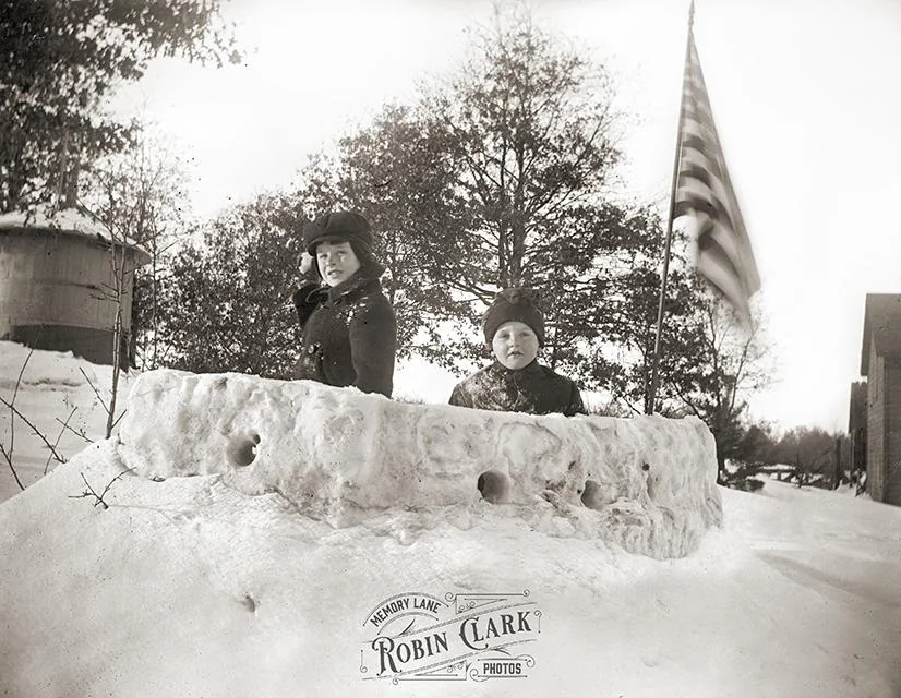 Two children in hats stand inside a semicircular snow fort with an American flag flying in the background.