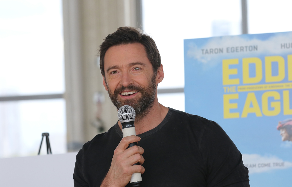 Hugh Jackman speaking into a microphone at an event with an “Eddie the Eagle” movie poster in the background.