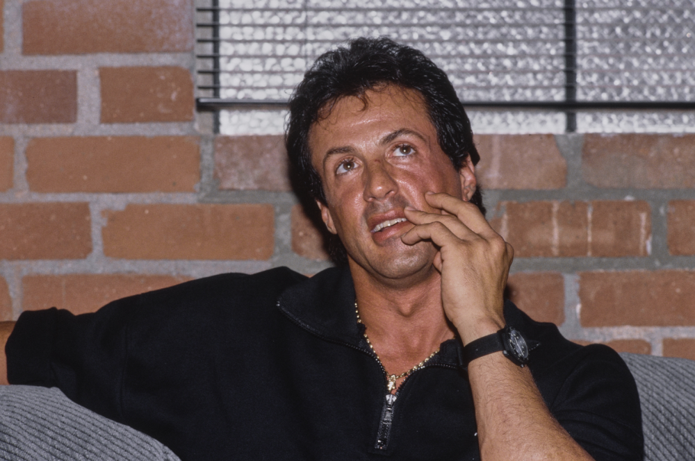 Sylvester Stallone resting his chin on his hand while sitting in front of a brick wall.