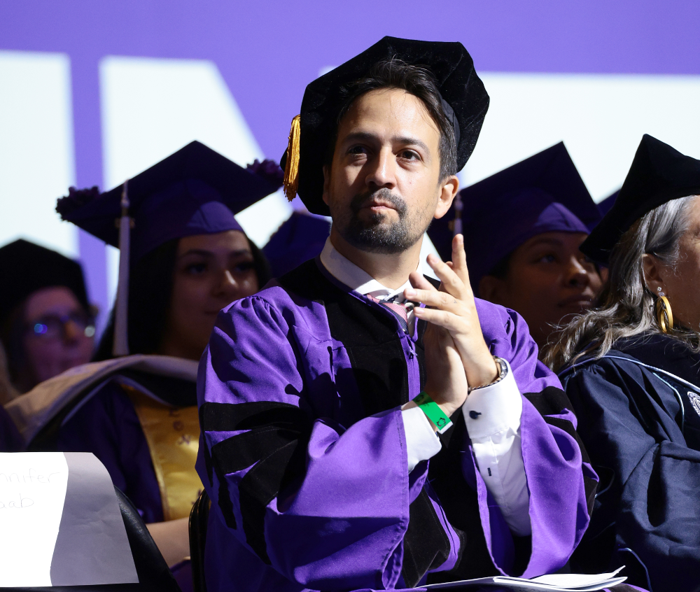 Lin-Manuel Miranda wearing a purple academic gown and cap while clapping at a graduation ceremony.