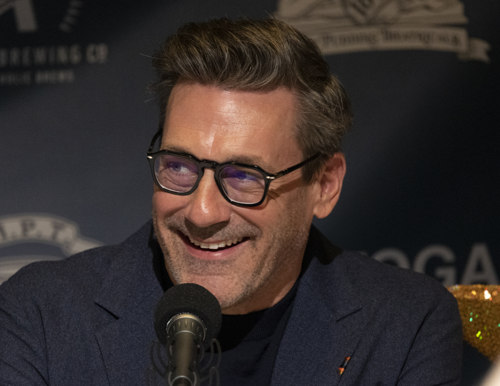 Jon Hamm wearing glasses and a dark blazer while speaking into a microphone.
