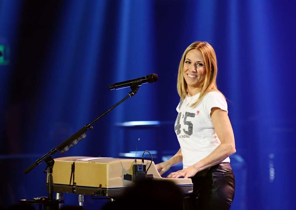 Sheryl Crow smiling while performing at a keyboard on stage.