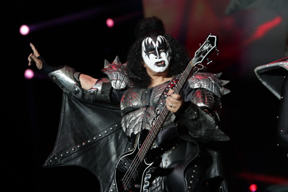 Gene Simmons of the band KISS performing on stage in full makeup and bat-wing stage armor while playing a bass guitar.
