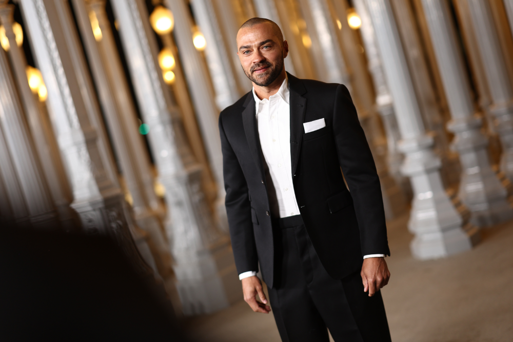 Jesse Williams standing in a suit and white shirt in front of a row of light posts.