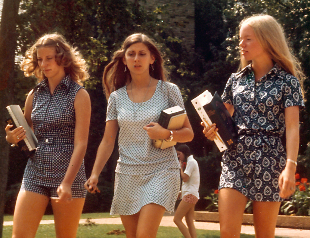 Three young women carrying books walk across a campus lawn.