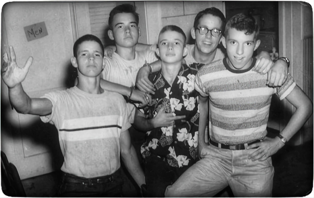 Five teenage boys pose for a black-and-white group photo, with one boy waving to the camera.