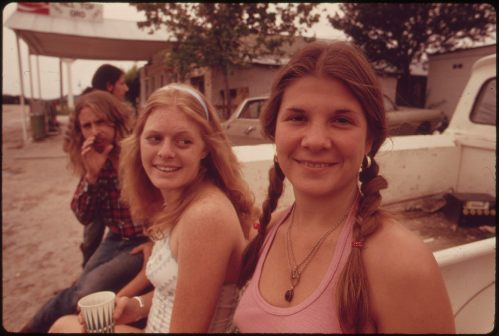 Two young women smiling while sitting on the back of a truck, with a young man smoking in the background.