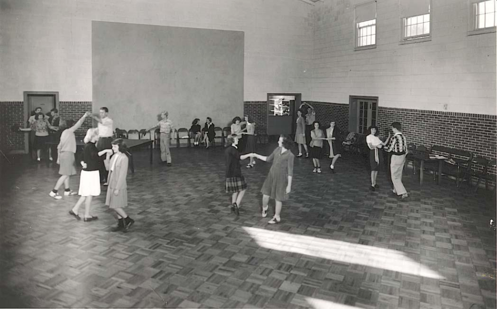 Several couples dance in a large hall with a checkered floor, while others stand along the walls.