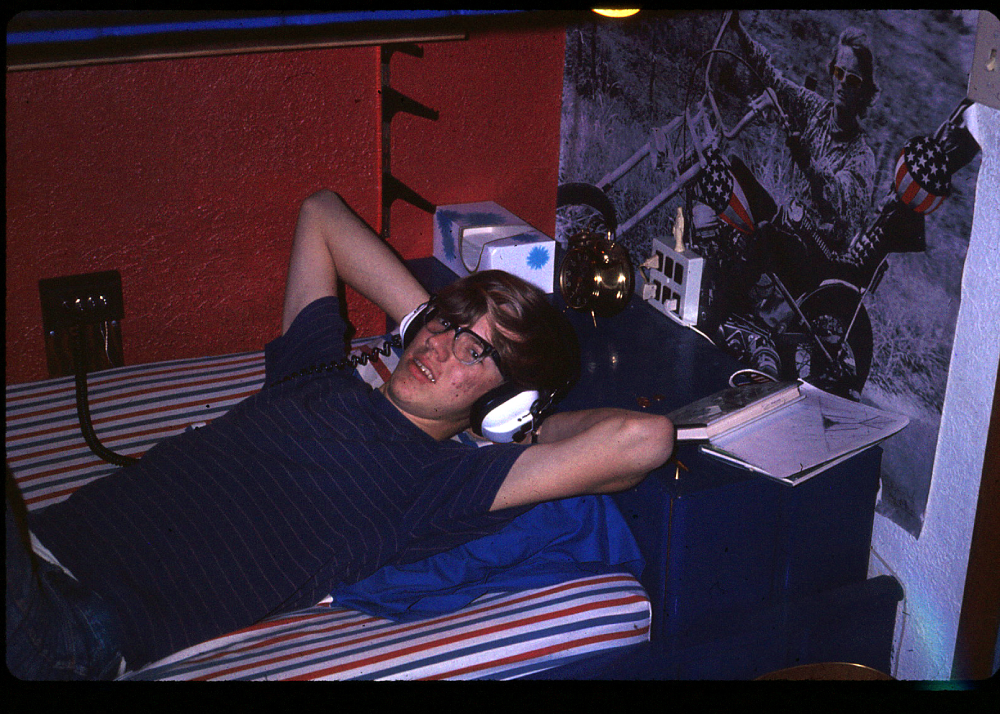 A young person with glasses and headphones lies on a bed, hands behind their head, next to a poster of a man on a motorcycle.