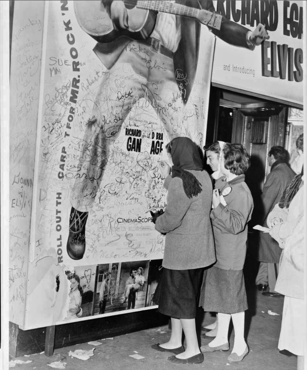 Two women standing on a sidewalk write their names on a large, graffiti-covered movie poster for the film “Love Me Tender” starring Elvis Presley.