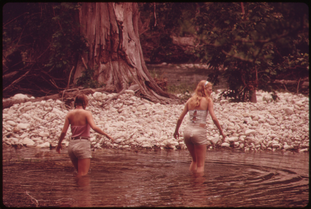 Two young women wade through a shallow river in front of a rocky bank and a large tree.