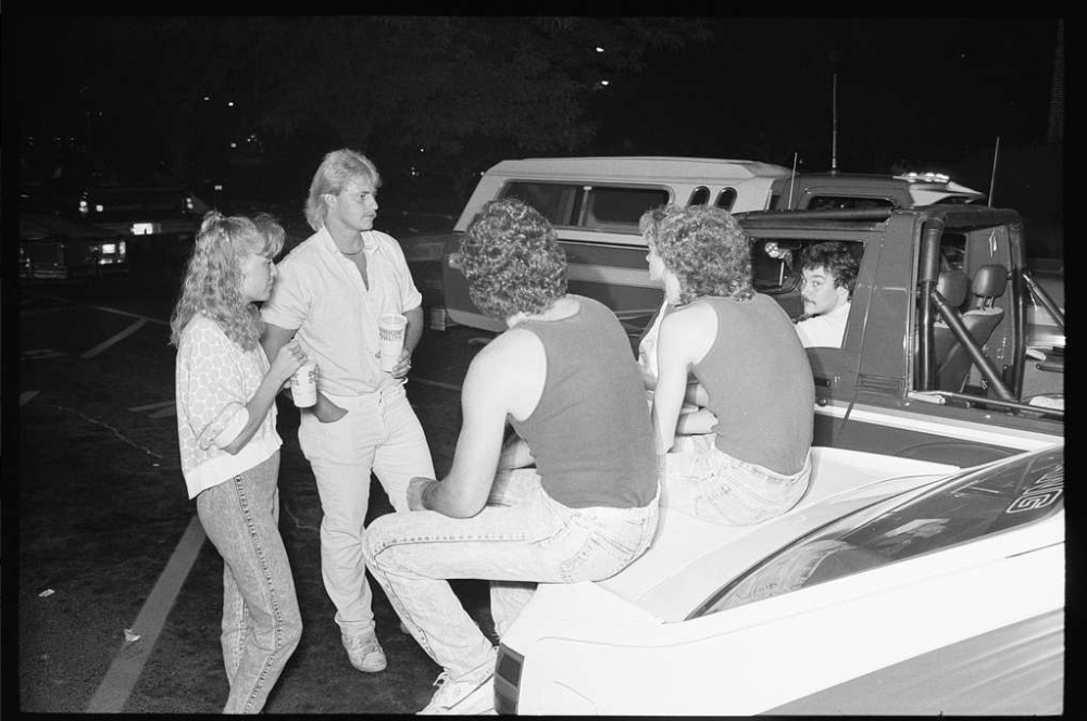 Four young people socialize in a parking lot at night, with two sitting on the tailgate of a pickup truck.