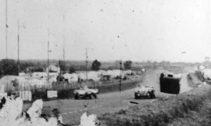 A grainy, black-and-white photograph shows a race car flipping over mid-air on a track with other vehicles nearby and a crowd in the background.