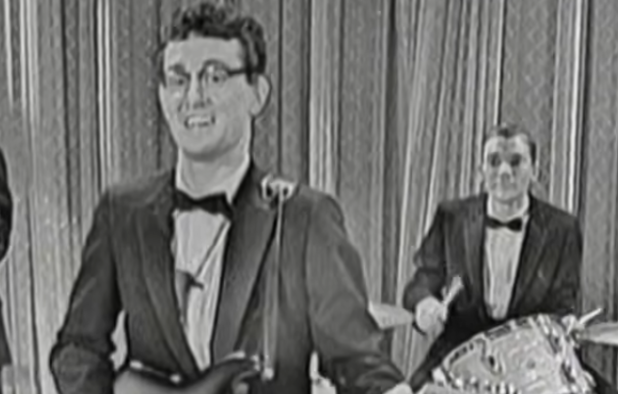Buddy Holly playing guitar and singing into a microphone, with a drummer performing in the background.