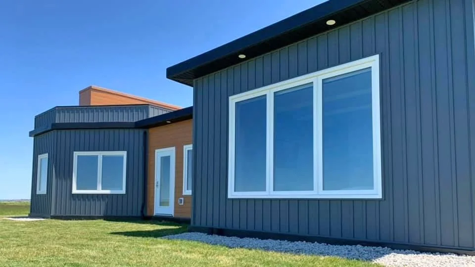 Modern home with dark blue vertical siding, large white-framed windows, and a glass door, set on a grassy lawn with a gravel border.