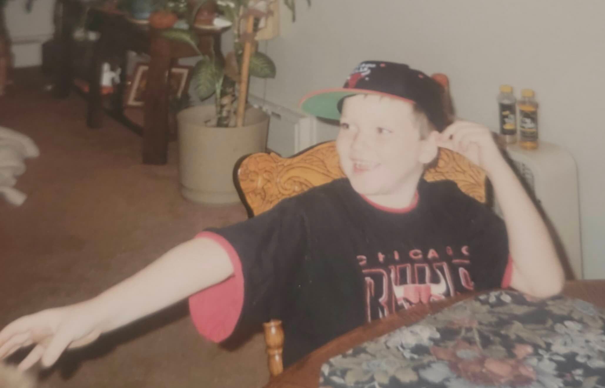 A young boy wearing a baseball cap and a Chicago Bulls t-shirt sits at a table and gestures with his arm.