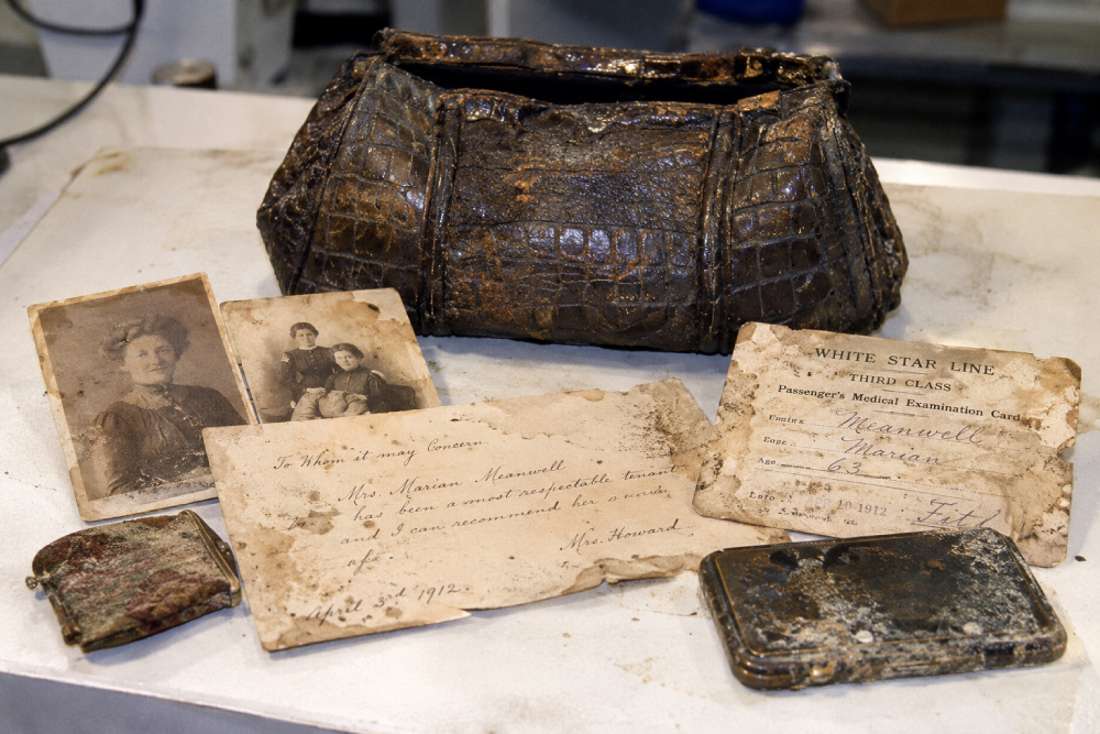A weathered leather handbag surrounded by two vintage photographs, a handwritten letter of recommendation for Mrs. Marian Meanwell dated April 1912, a White Star Line medical examination card, a small coin purse, and a metal case.