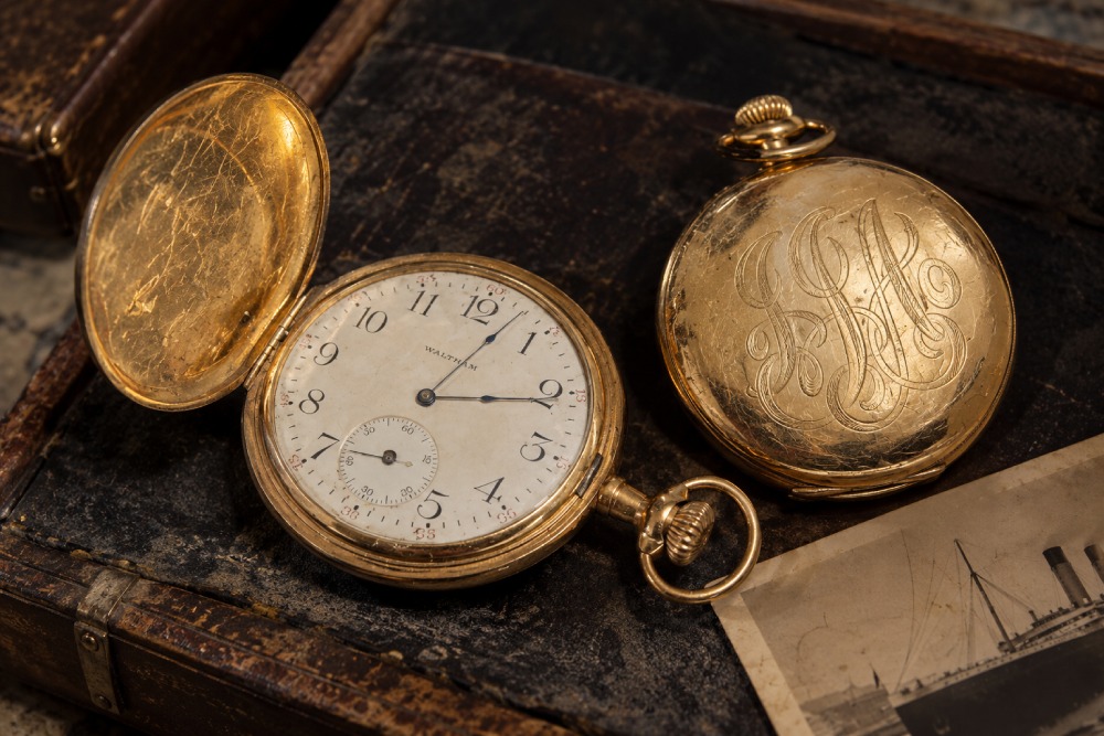 Two gold pocket watches, one open showing a Waltham watch face and one closed with an engraved monogram, resting on a dark antique case next to a vintage photograph.